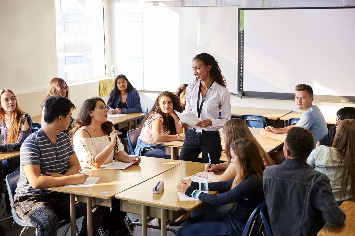 Female High School Teacher Standing By Student Table Teaching Lesson